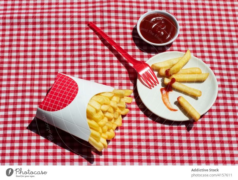 French fries packet near plate with potatoes soaked in ketchup american culture condiment potato chips unhealthy eating red background white frites take out