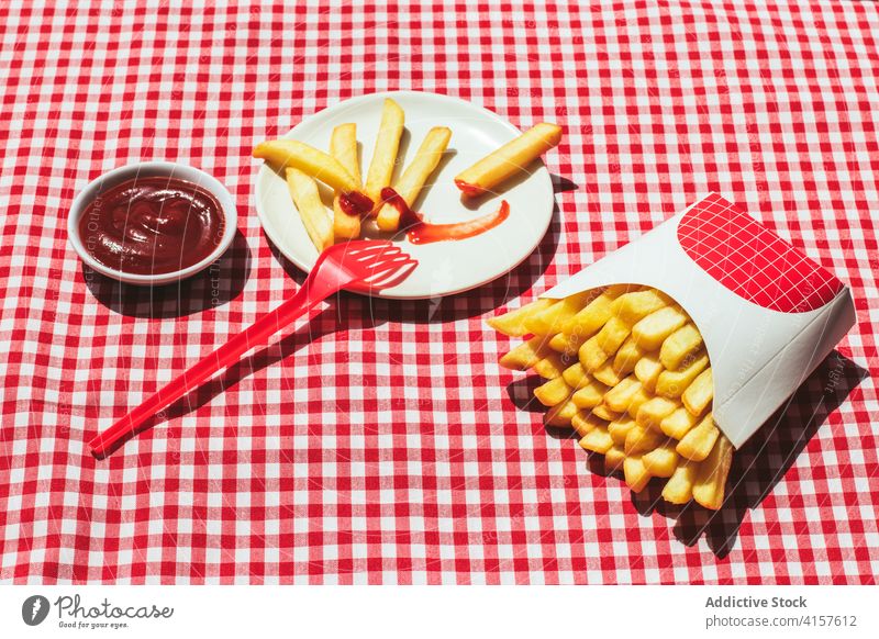 French fries packet near plate with potatoes soaked in ketchup american culture condiment potato chips unhealthy eating red background white frites take out