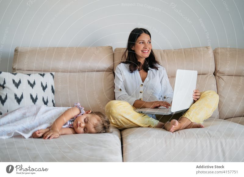 Smiling woman working on the sofa with the laptop while a toddler is sleeping on a side maternity responsibility tenderness dreaming motherhood newborn asleep