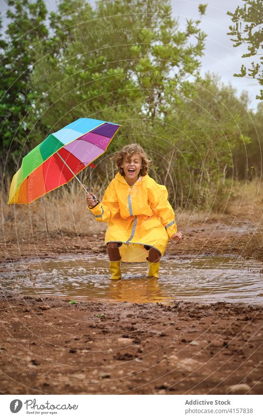Child in a raincoat with umbrella crouched down playing and laughing in a puddle schoolboy profile shower attitude rainbow curly gesture enjoyment hold pouring