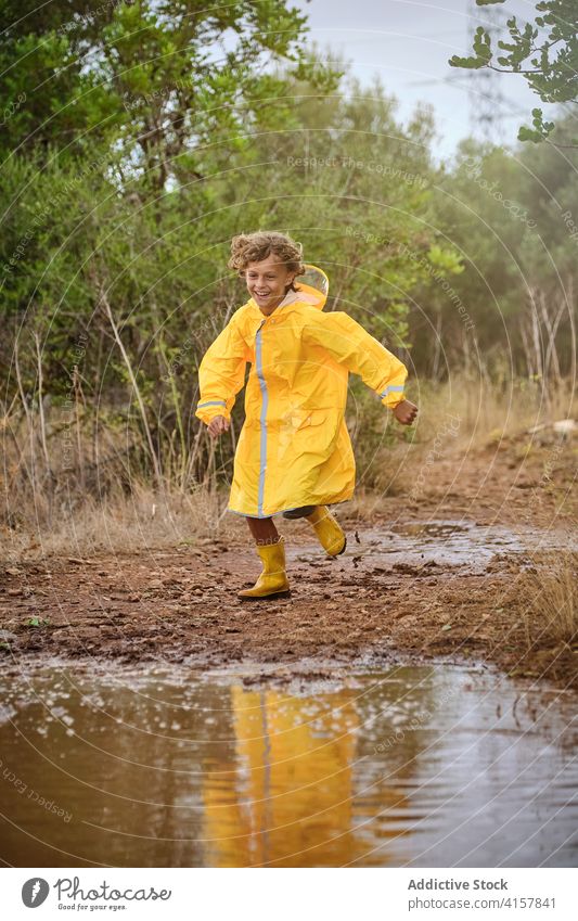 Boy in a raincoat and rain boots running and playing along a path full of puddles laugh schoolboy shower attitude curly enjoyment speed pouring wet weather