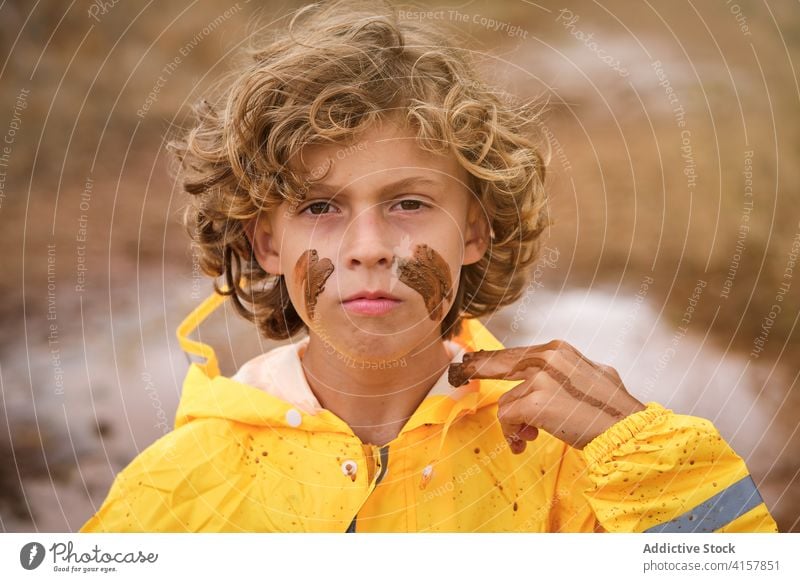 Kid in raincoat drawing marks on his face with the mud facing the camera seriously pride infant messy playful fingers ethnic storm curly makeup joke youth wet