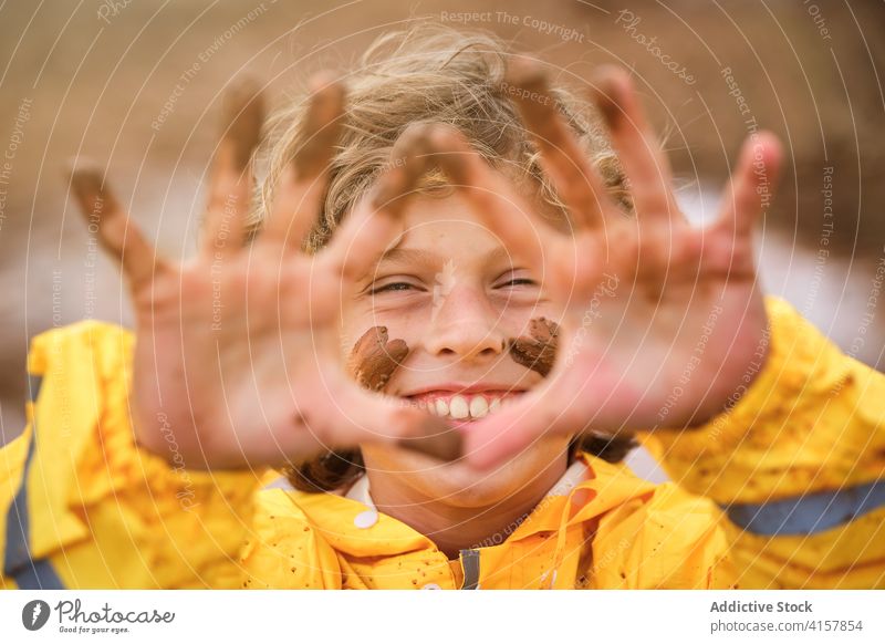 Selective focus on the muddy and happy face of a child that is showing the dirty hands in raincoat outdoors joke playful infant happiness messy fingers mark