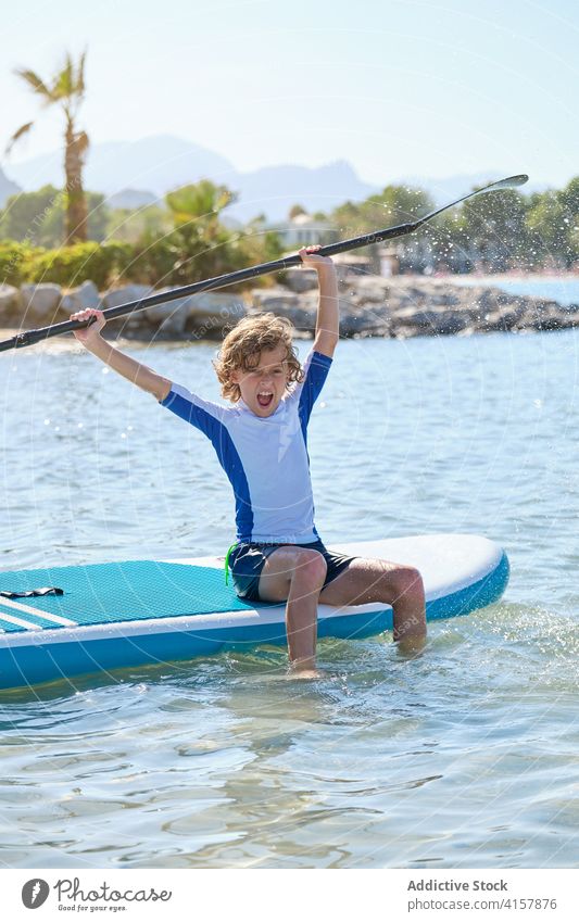 Boy with curly hair sitting on a paddle surfboard and playing with the paddle splashing withe the water vitality gesture arms raise balance enjoy recreation