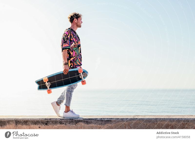 Stylish man with skateboard walking on seafront skater embankment hipster style trendy activity summer young male lifestyle modern millennial funky active deck