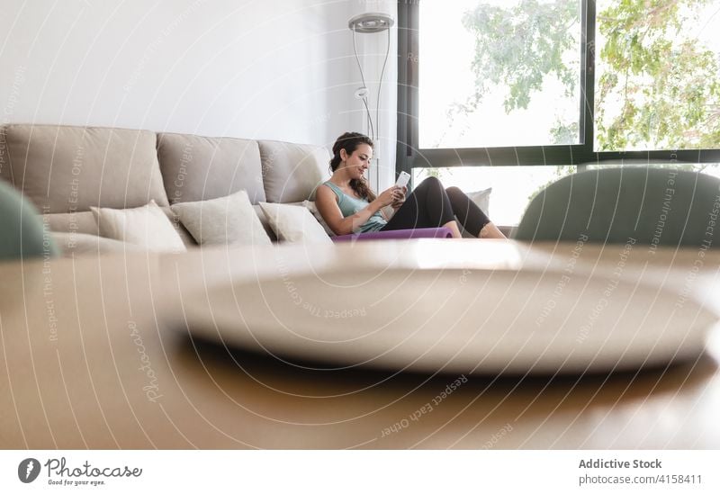 Tranquil woman relaxing on sofa with smartphone and yoga mat browsing home lying using weekend female sportswear activewear living room couch soft modern
