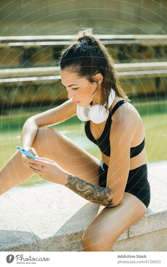 Sportive woman with smartphone and headphones resting after training on street sportswoman using browsing gadget athlete workout tattoo mobile device relax