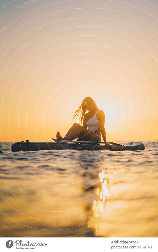 Carefree woman on paddleboard in sea relax sunset sup surfboard slim sundown female swimsuit ocean summer sit rest enjoy holiday nature fit sky tranquil evening