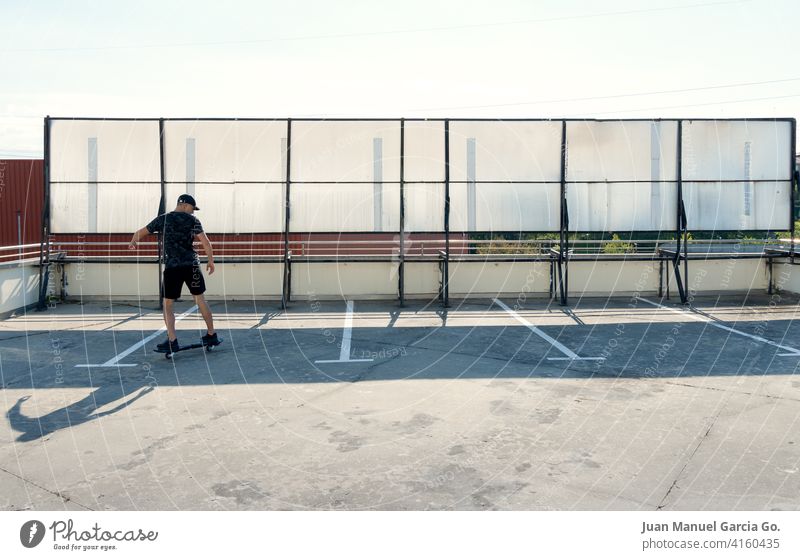 Teenager dressed in black and cap skateboarding in industrial zone teenager parking lot abandoned layered haircut solo rooftop seriousness clear sky shorts