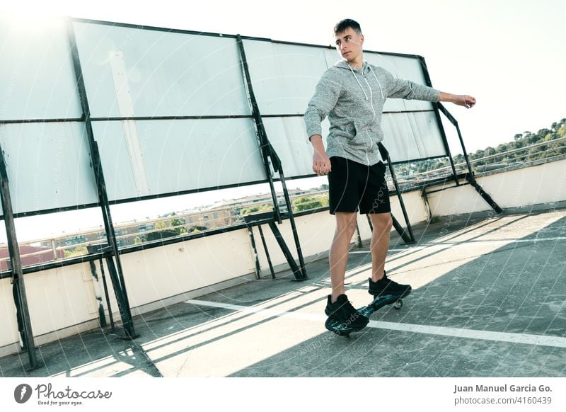 Rider on waveboard plays in abandoned parking lot teenager sweatshirt layered haircut alone rooftop seriousness clear sky shorts sport young leisure game