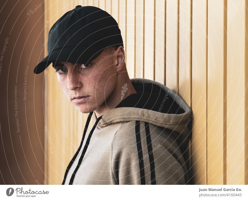 Teenager with cap in foreground looks at camera teenager black sweatshirt khaki serious intense puberty pimples acne bald door industrial zone tracksuit