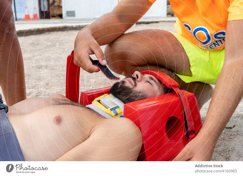 Crop lifeguard rescuing patient in neck brace on beach rescue aid training practice eyes closed immobilizer medical emergency head block provide help seashore