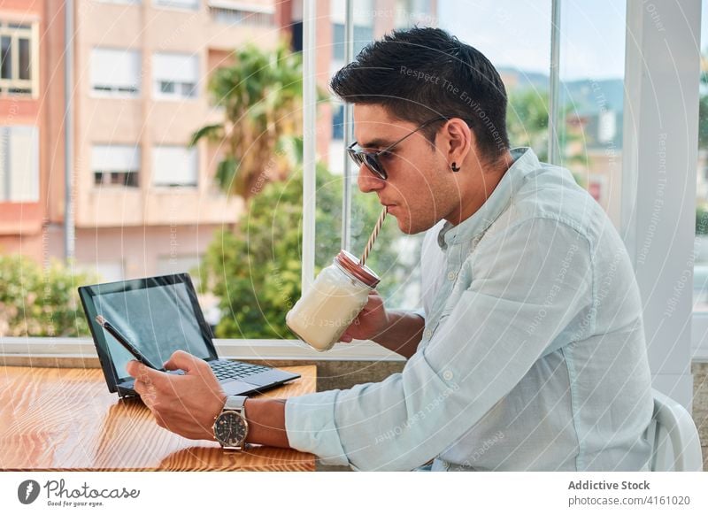 Focused young man drinking beverage while working distantly using laptop and smartphone milkshake freelance focus online connection internet male style