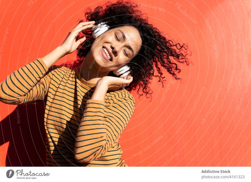 Cheerful ethnic woman listening to music in headphones headset enjoy eyes closed trendy appearance touch face bright song dreamy afro hairstyle happy cheerful