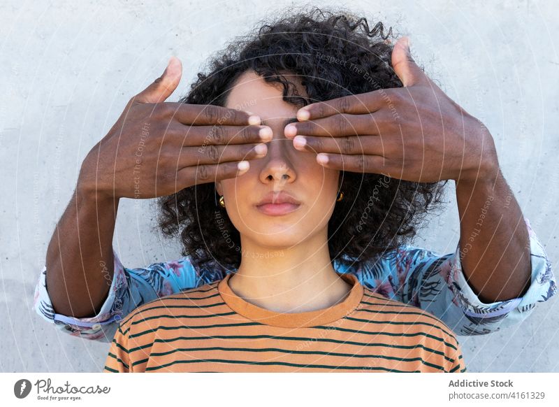 Anonymous partner covering eyes of black woman near cement wall cover eyes friend focus stylish appearance curly hair hairstyle attentive portrait trendy modern