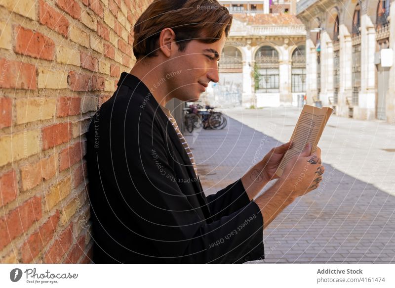 Crop smiling trendy tattooed man reading book on pavement appearance literature smile brick wall town stylish apparel street content wear old building shabby