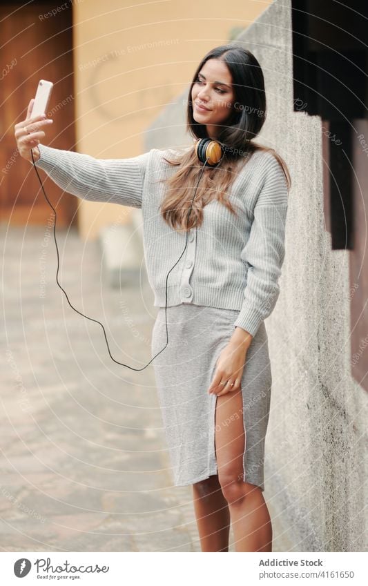 Charming woman with smartphone and headphones on street taking selfie music style connect tranquil casual rest melody listen female city urban audio relax