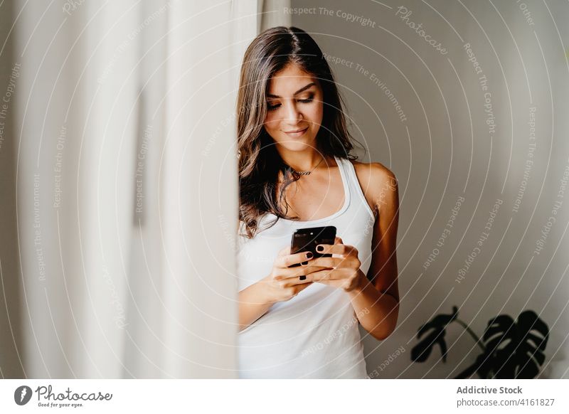 Young woman on smartphone standing near window in morning using home browsing gadget millennial female lifestyle bedroom device internet online social media