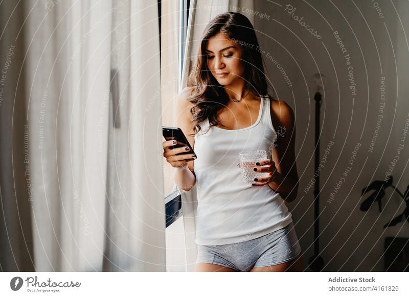 Young woman with glass of water and smartphone standing near window in morning drink using home browsing gadget millennial female lifestyle bedroom device