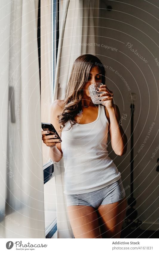 Young woman with glass of water and smartphone standing near window in morning drink using home browsing gadget millennial female lifestyle bedroom device