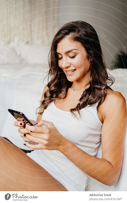 Happy woman on smartphone and leaning on bed charming speak cellphone smile bedroom content communicate gadget device using brunette young call female cheerful