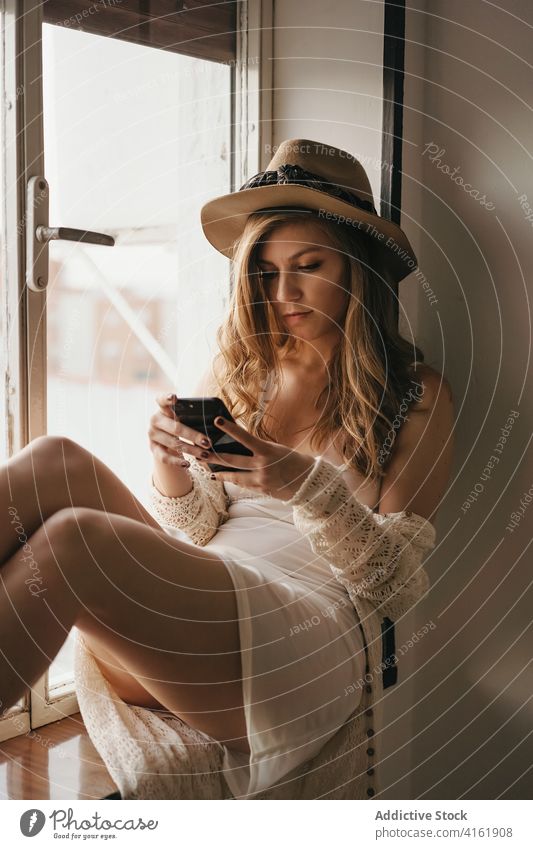 Stylish seductive woman chatting on smartphone near window stylish apparel sensual feminine using gadget boho device sensitive windowsill home straw hat trendy