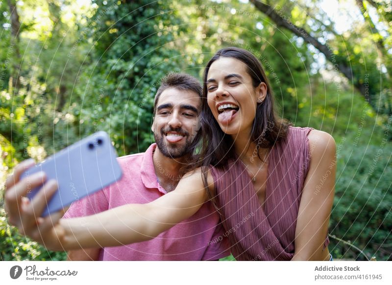 Cheerful ethnic couple taking selfie in park tongue happy smartphone cuddle date romantic love together relationship gadget mobile girlfriend boy young casual