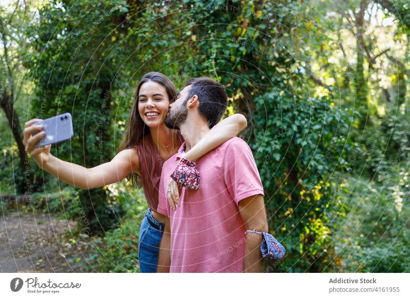 Cheerful ethnic couple taking selfie in park kiss happy smartphone cuddle date romantic love together relationship gadget mobile girlfriend boy young casual