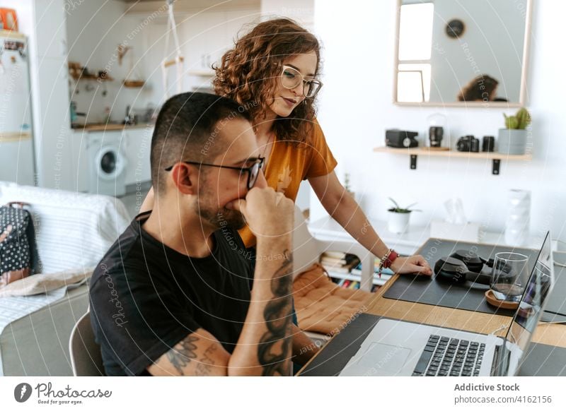 Concentrated young diverse couple working on laptop at home remote support teamwork freelance concentrate using device gadget boyfriend girlfriend eyeglasses