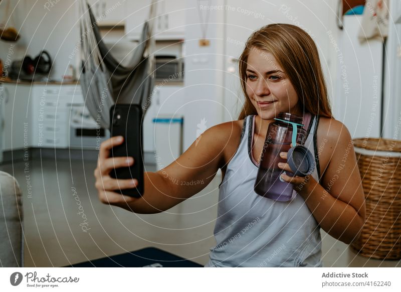 Content sportswoman with water bottle taking selfie on smartphone vitality energy training workout smile content hydrate beverage practice at home activity