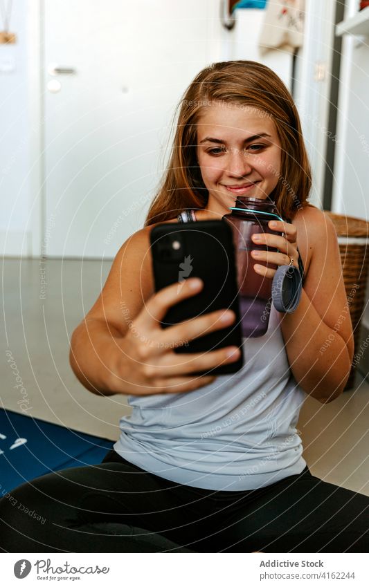 Content sportswoman with water bottle taking selfie on smartphone vitality energy training workout smile content hydrate beverage practice at home activity