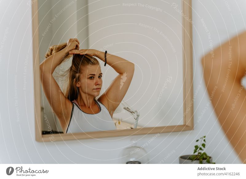 Calm woman doing ponytail against mirror hairstyle hairdo calm reflection fix routine concentrate serious casual young focus attractive trendy slim stand modern