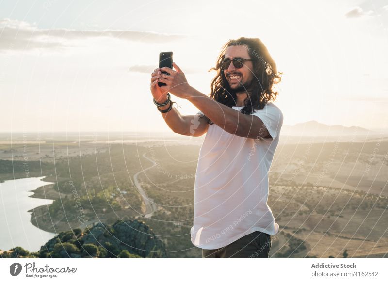 Cheerful man taking selfie against sunny lush valley traveler cheerful toothy smile smartphone nature excited moment gadget hilltop wanderlust amazing viewpoint