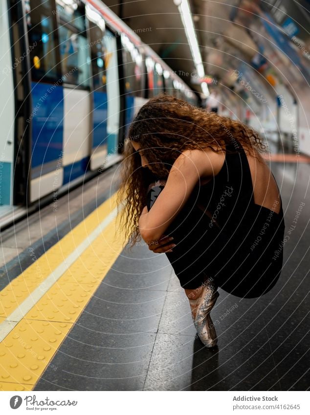 Anonymous young ballerina sitting on tiptoe on metro platform woman embracing knee haunch subway station perform melancholy grace style train female long hair