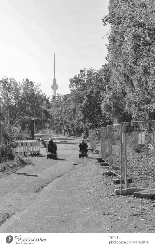 two men drive in an e-wheelchair through the Mauerpark towards the television tower wall park Television tower Prenzlauer Berg bnw b/w Berlin Wheelchair