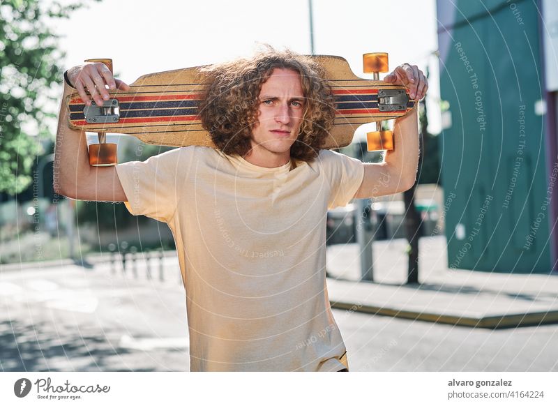 young man with curly hair looking at camera with his longboard or skeateboard on his shoulders in the middle of the street che skateboard attractive person