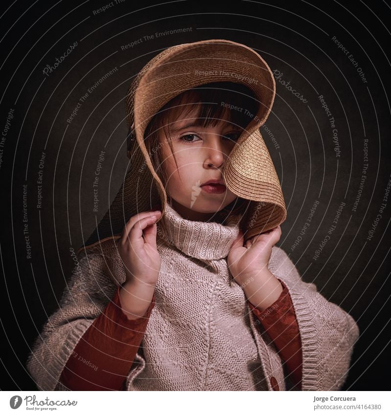 4-year-old Caucasian girl with hat and a cloth poncho. she poses stylish and elegant holding the hat with her hands. square format adorable alone attractive
