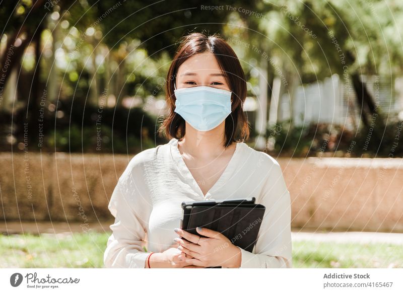 Young ethnic woman working remotely on tablet in park attentive new normal coronavirus job busy gadget using browsing female young asian formal mask covid 19