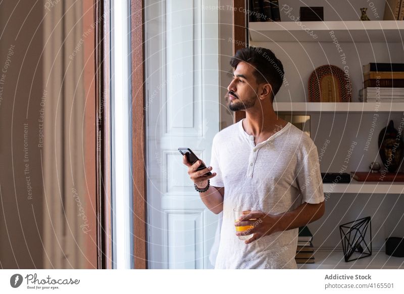 Young man with glass of juice browsing smartphone at home morning window using drink gadget lifestyle young male beard modern beverage mobile connection