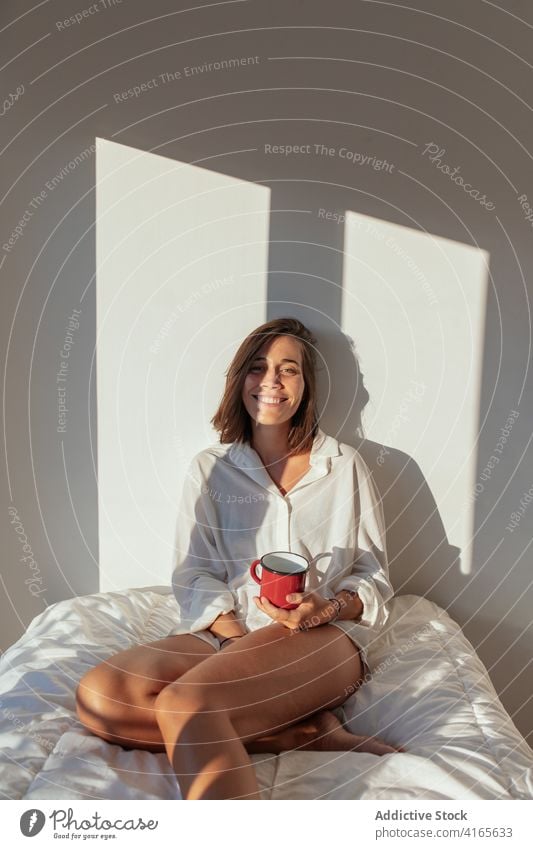 Cheerful young woman drinking coffee in bed morning smile happy sensual comfort allure cozy joy female white shirt body barefoot home beverage sunny relax rest