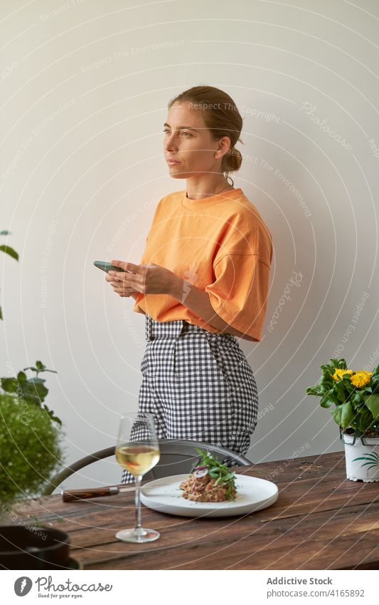 Calm woman using smartphone at home during lunch steak tartare serve table wine relax female cellphone browsing beverage gadget device pensive plate glass drink