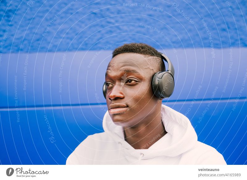 Young afro American black man listening music with wireless headphones while wearing a white sweatshirt, looking to the side young african blue street