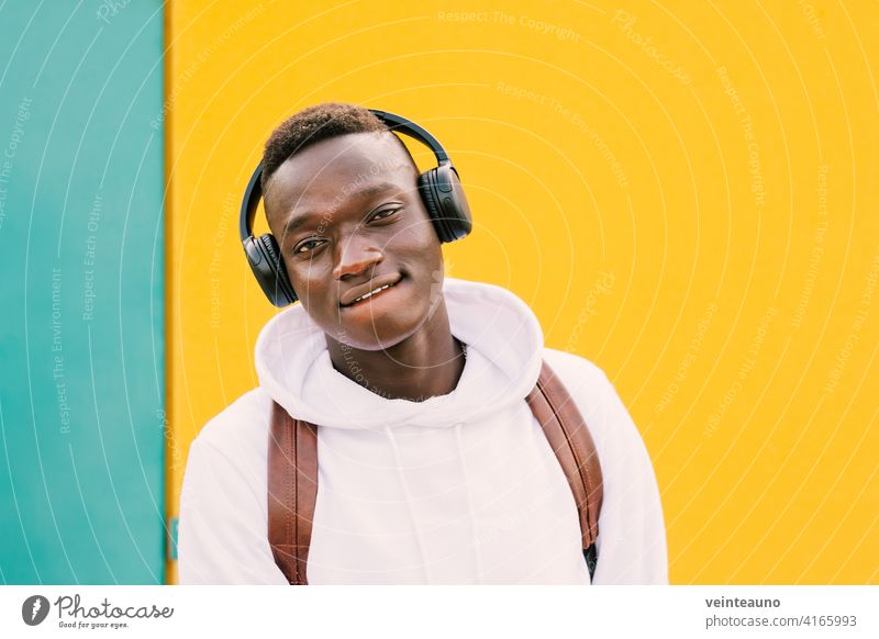 Young afro American black man listening music with wireless headphones while wearing a white sweatshirt and a backpack looking to camera on a green and yellow wall background