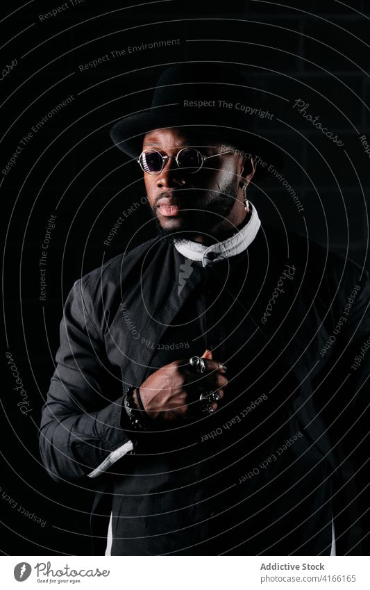 Calm stylish man in dark studio style black cloth ring hat apparel trendy sunglasses male ethnic african american round shape serious cool model stand accessory