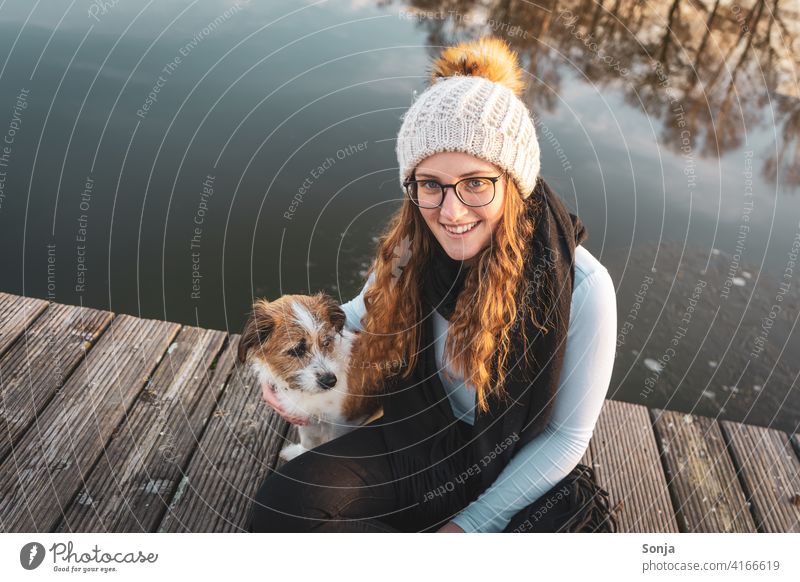 Young woman with glasses sits with a small dog on a wooden jetty by the lake Woman youthful Laughter Eyeglasses Dog Small Sit wooden walkway Lake Winter Cold