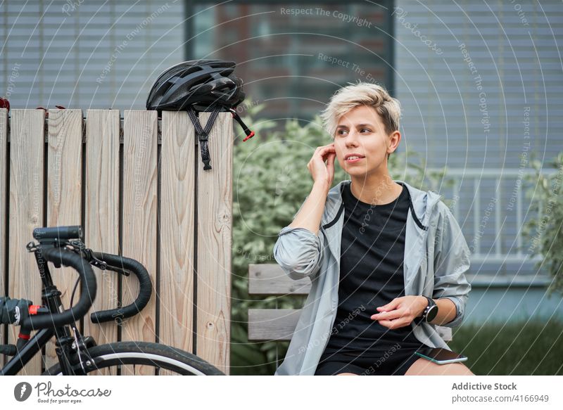 Female courier talking on phone while sitting on bench next to bicycle on background of building Cyclist city day woman young sports riding summer helmet