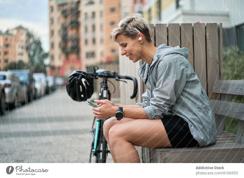 Side view of happy woman courier with phone in his hands sits on bench next to bicycle on background of building Cyclist city day young sports riding summer