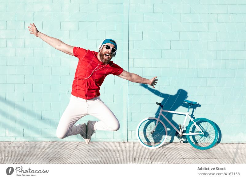 Cheerful young man jumping on street near modern bike and blue building outstretch cheerful expressive scream bicycle trendy urban happy male hipster outfit