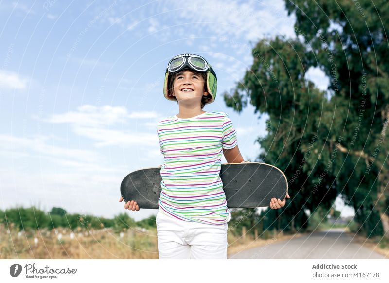 Happy dreamy boy with skateboard on roadway child goggles happy countryside stylish apparel cloudy blue sky childhood kid positive smile enjoy stripe wear