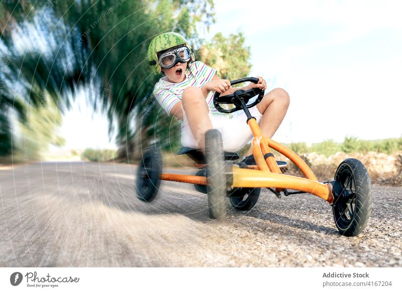 Excited boy riding pedal car on road ride amazed recreation having fun activity excited goggles childhood trendy wear kid go kart transport roadway asphalt